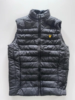 Svart dunväst, Lyle & Scott - Snygg svart dunväst med quiltad design och hög krage. Västen har en gul broderad logga på bröstet och dragkedja framtill. Perfekt för lager-på-lager och håller dig varm utan att bli för tung. Passar till de flesta outfits och är riktigt stilren.