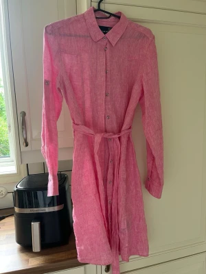 Rosa linneskjortklänning från Pelle P - Säljer en rosa skjortklänning från Pelle P i linne med knappar framtill, klassisk krage och avtagbart knytskärp i midjan. Klänningen har långa ärmar som kan vikas upp och fästas med knapp. Perfekt för sommarens alla tillfällen!