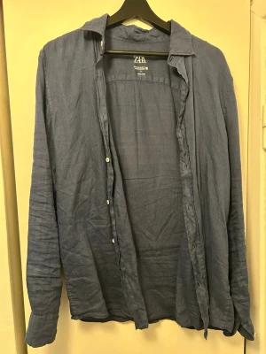 Blå linneskjorta från Zara - Säljer en stilren blå skjorta från Zara i tunt och luftigt linne. Skjortan har klassisk krage, långa ärmar och knappar framtill. Perfekt för dig som gillar en avslappnad men snygg look.