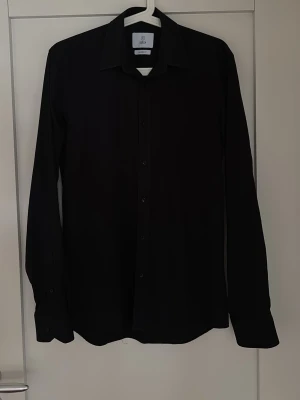 Bläck svart skjorta - Stilren svart skjorta med lång ärm och knappar framtill. Skjortan har klassisk krage och är perfekt för dig som gillar en enkel och clean look. Passar till många olika outfits och är ett måste i garderoben.                         