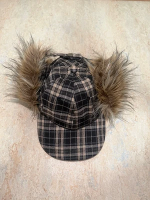 Rutig keps med fuskpälsöron - Säljer en unik keps i svart och beige rutigt tyg med stora, fluffiga fuskpälsöron i brunt. Kepsen har böjd skärm och coolt vinterinspirerat utseende. Perfekt för dig som vill sticka ut med din stil och gillar accessoarer med attityd.