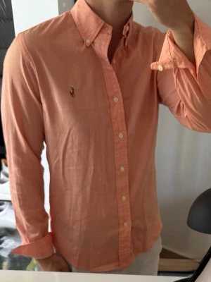 Persikofärgad skjorta från Ralph Lauren - Säljer en persikofärgad skjorta från Ralph Lauren med klassisk button-down krage och broderad logga på bröstet. Skjortan har långa ärmar, vita knappar och är tillverkad i bomull. Perfekt för dig som vill ha en stilren och fräsch look.