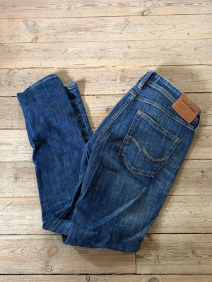 Jack&Jones Jeans - Säljer ett par klassiska blå jeans från Jack&Jones med raka ben och femficksmodell. Jeansen har snygga slitningar och kontrastsömmar samt patch i läder bak i midjan. Tillverkade i bomull och perfekta för en avslappnad stil.