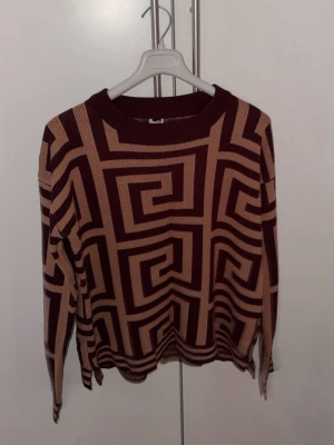 Brun och beige stickad tröja med mönster - Säljer en stickad tröja i brunt och beige med ett grafiskt, labyrintliknande mönster. Tröjan har rund halsringning och långa ärmar. Perfekt för dig som gillar unika och trendiga plagg med lite extra edge.