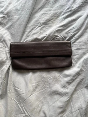 Mörkbrun kuvertväska i skinn - Stilren mörkbrun kuvertväska i mjukt skinn med enkel design. Väskan har ett magnetlås i guldton och en vikbar öppning. Perfekt för att bära det viktigaste och ger en klassisk touch till din outfit.