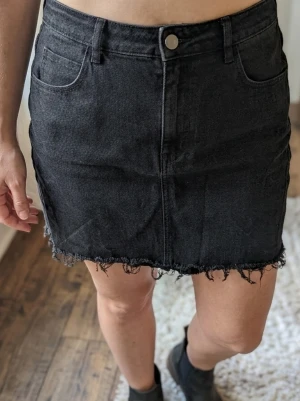 Svart jeanskjol med fransad kant - Säljer en svart jeanskjol med fransad nederkant och klassisk femficksdesign. Kjolen har normal passform och stängs med knapp och dragkedja framtill. Perfekt till en avslappnad och trendig look.