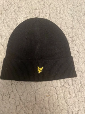 Svart ribbad mössa med gul logga - Säljer en svart ribbstickad mössa med uppvikt kant och en gul broderad logga framtill. Klassisk och enkel design som passar till det mesta. Perfekt för kalla dagar och enkel att matcha med olika outfits.