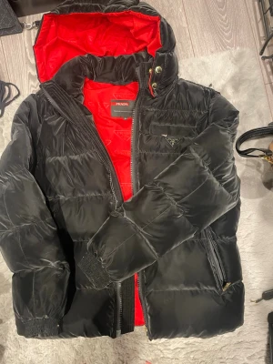 Svart dunjacka från Prada med röd insida - Snygg svart dunjacka från Prada med röd insida och huva. Jackan har dragkedja, tryckknappar i guld och Pradas ikoniska trekantslogga på bröstet. Perfekt för kalla vinterdagar och riktigt stilren look.