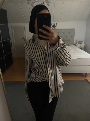 Randig beige  skjorta med svarta ränder - Säljer en snygg vit skjorta med svarta vertikala ränder. Skjortan har klassisk krage, knäppning framtill och långa ärmar. Passformen är normal och materialet känns lätt och bekvämt. Perfekt för en stilren look.