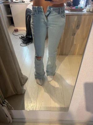 Ljusblå bootcut jeans med hål - Säljer ett par ljusblå bootcut jeans med låg midja och ett snyggt hål på ena knät. Jeansen har klassisk femficksdesign och är tillverkade i denim med lite stretch. Perfekta för en avslappnad och trendig look. Har två små hål på sidan men inget som syns mycket