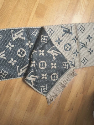 Beige och grå Louis Vuitton halsduk - Säljer en lyxig halsduk från Louis Vuitton i beige och grått med ikoniskt monogrammönster. Halsduken är vävd och har fransar i ena änden. Perfekt accessoar för att lyfta din outfit med exklusiv känsla.
