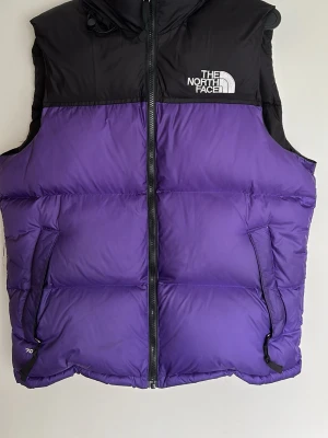 Lila och svart dunväst The North Face - Snygg dunväst från The North Face i lila och svart med dragkedja framtill och två fickor. Västen har hög krage och logga på bröstet. Perfekt för lager-på-lager och håller dig varm med sin fluffiga dunfyllning.