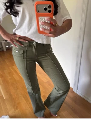 Olivgröna bootcut jeans med vita knappar - Snygga olivgröna jeans med bootcut passform och markerade sömmar. Jeansen har vita knappar och fickdetaljer framtill som ger en cool retrovibe. Perfekta för dig som vill sticka ut lite extra med din stil.