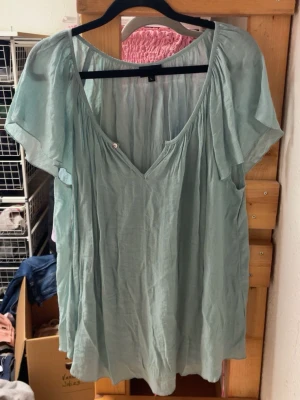 Ljusgrön luftig blus med v-ringning - En ljusgrön blus i ett tunt och luftigt tyg med avslappnad passform. Blusen har korta ärmar och djup v-ringning framtill. Perfekt för varma dagar och enkel att styla till olika outfits. Materialet ser ut att vara viskos eller bomull.