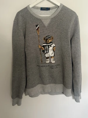 Grå sweatshirt med Polo Bear - Grå sweatshirt från Polo Ralph Lauren med det ikoniska Polo Bear-motivet framtill. Tröjan har rund halsringning, ribbade muddar och nederkant samt en avslappnad passform. Perfekt för en chill och stilren look.