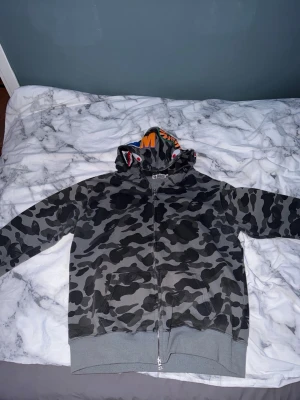 Grå camo hoodie från A Bathing Ape - Säljer en grå camo zip hoodie från A Bathing Ape med ikonisk hajdesign på huvan och färgglada bokstäver. Hoodien har dragkedja, ribbade muddar och är tillverkad i mjuk bomull. Perfekt för dig som gillar streetwear och vill sticka ut. Riktig fet. LITEN I STORLEK SÅ PASSAR L. PRIS GÅR ATT DISKUTERA