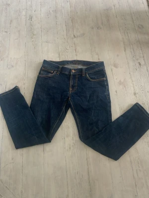 Mörkblå jeans från Nudie Jeans - Säljer ett par klassiska mörkblå jeans från Nudie Jeans med raka ben och orange kontrastsömmar. Jeansen har fem fickor och är tillverkade i slitstark denim. Perfekta för en avslappnad och stilren look.