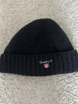 Svart ribbad mössa från GANT - Säljer en svart ribbstickad mössa från GANT med uppvikt kant och broderad logga framtill. Klassisk och enkel design som passar till det mesta. Perfekt för hösten och vintern som kommer snart! Säljer för 250 kan möjligtvis gå med på lägre!