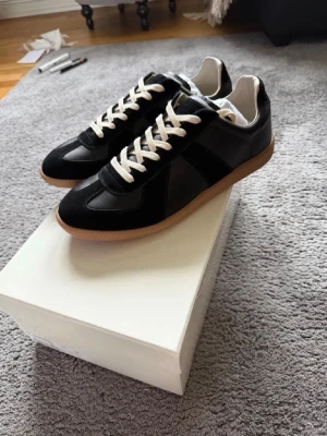 Maison margiella gats svart - Stilrena svarta sneakers med vita skosnören och beige gummisula. Ovandelen är i en mix av läder och mocka för en schysst kontrast. Klassisk låg modell som passar till det mesta och ger en clean look.