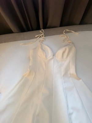 Vit långklänning med knytband - Elegant vit långklänning i ett slätt, glansigt material med tunna axelband som knyts på axlarna. Klänningen har en figurnära överdel med markerade kupor och en lätt utsvängd kjol. Perfekt för sommarens alla tillfällen.