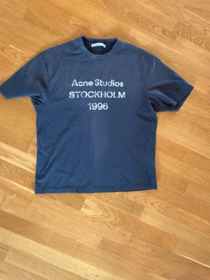 Blå Acne Studios t-shirt Stockholm - Snygg blå t-shirt från Acne Studios med tryck 'Acne Studios STOCKHOLM 1996' på bröstet. Klassisk passform och rund hals. Perfekt till jeans eller shorts för en clean och enkel look. Det finns inga skönhets skador på tröjan.