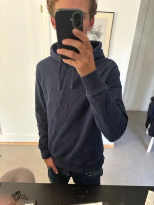 Mörkblå hoodie - En stilren mörkblå hoodie med huva och dragsnören. Hoodien har en klassisk passform, ribbade muddar vid ärmslut och nederkant samt är tillverkad i mjukt bomullsliknande material. Perfekt för en avslappnad och trendig look.