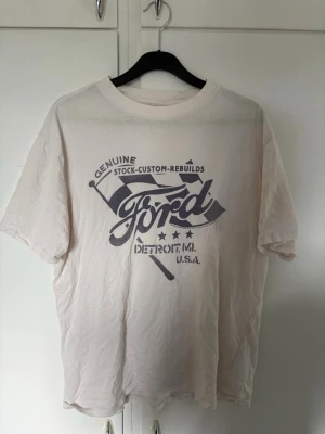Vit t-shirt från Abercrombie - Säljer en vit t-shirt med stort retroinspirerat Ford-tryck i svart på bröstet. T-shirten har rund hals och korta ärmar. Trycket har texten 'Genuine Stock-Custom-Rebuilds Ford Detroit, MI, U.S.A.' och flaggdetaljer. Perfekt för dig som gillar bilar och vintage-stil. Bra skick 