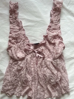 Rosa spetstopp med volanger - Söt rosa topp från Gina Tricot i transparent spets med volangdetaljer och breda axelband. Dekorerad med en liten rosett framtill och har en romantisk, feminin look. Perfekt för dig som gillar mjuka pasteller och detaljer.
