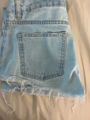 Ljusblå jeansshorts med fransig kant - Säljer ett par ljusblå jeansshorts i klassisk femficksmodell. Shortsen har rå, fransig kant nertill och knappar i gylfen. Perfekta för sommaren och ger en avslappnad vibe till din outfit.