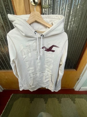 Hollister hoodie - Vit hoodie från Hollister med stor logga och fågeltryck i vinrött på bröstet. Tillverkad i 60% bomull och 40% polyester. Klassisk känguruficka framtill och justerbar dragsko i huvan. Perfekt för en avslappnad och chill stil.