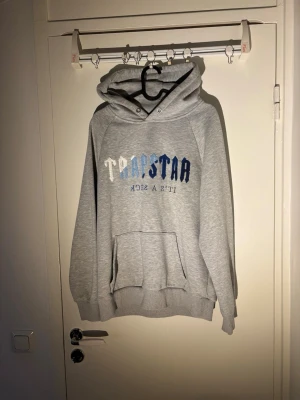 Trapstar hoodie - Säljer en grå hoodie från Trapstar med stor magficka och tryck i vitt och blått på bröstet. Hoodien har huva med dragsko och ribbade muddar vid ärmar och nederkant. Perfekt för en chill och avslappnad stil.