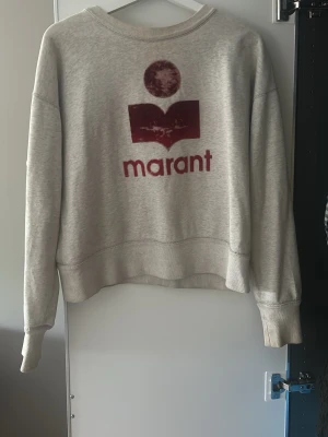 Isabel Marant Grå sweatshirt  - Säljer en grå sweatshirt från Marant med stor röd logga och text framtill. Tröjan har rund halsringning, långa ärmar och ribbade muddar vid ärmslut och nederkant. Perfekt för en avslappnad och trendig look.