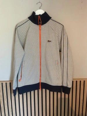Houndstooth ziptröja från Lacoste - Snygg ziptröja från Lacoste med klassiskt hundtandsmönster i svart, vitt och beige. Tröjan har orange dragkedja, hög krage och muddar i mörkblått. Ikoniska Lacoste-loggan på bröstet. Perfekt för dig som gillar retrostil och streetwear.
