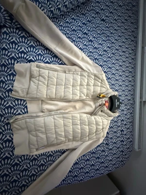Beige quiltad jacka med dragkedja - Snygg beige jacka med quiltade partier framtill och på axlarna. Jackan har dragkedja, lång ärm och en stilren, enkel design. Perfekt för dig som gillar en clean och modern look.