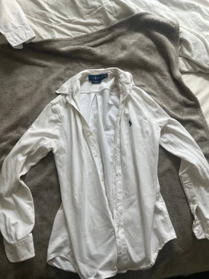 Vit skjorta från Ralph Lauren - Klassisk vit skjorta från Ralph Lauren med blå broderad logga på bröstet. Skjortan har lång ärm, knappar framtill och en tidlös krage. Tillverkad i mjuk bomull för en bekväm känsla och snygg passform.  Och den e i linne skjorta