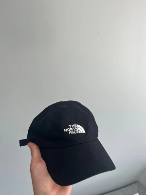Svart keps från The North Face - Snygg svart keps från The North Face med klassisk böjd skärm och justerbart band bak. Kepsen har broderad vit logga framtill och är tillverkad i ett mjukt tyg som känns bekvämt på huvudet. Perfekt för en clean och sportig stil.