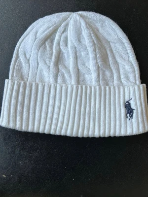 Vit kabelstickad mössa från Polo Ralph Lauren - Säljer en vit kabelstickad mössa från Polo Ralph Lauren med uppvikt kant och klassisk marinblå logga broderad framtill. Mössan är gjord i mjukt material och har en stilren look som passar perfekt till kalla dagar.