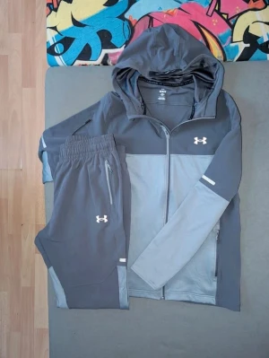 Grå träningsset från Under Armour - Snyggt träningsset från Under Armour i två nyanser av grått. Setet består av en zip-hoodie med huva och två fickor samt matchande byxor med resår i midjan och dragkedjor vid fickorna. Perfekt för gymmet eller löprundan. Materialet är mjukt och stretchigt för maximal komfort.