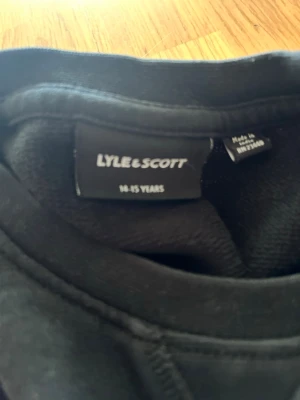 Svart sweatshirt från Lyle & Scott - Klassisk svart sweatshirt från Lyle & Scott med rund halsringning och diskret gul logga på bröstet. Tröjan har långa ärmar och ribbade muddar vid ärmslut och nederkant. Perfekt för en avslappnad och stilren look.