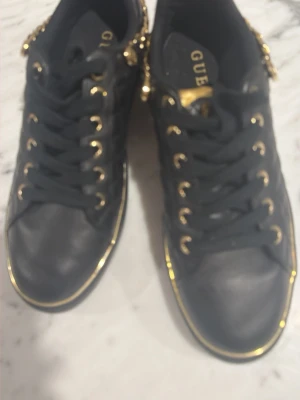 Svarta sneakers med gulddetaljer Guess - Snygga svarta sneakers från Guess med guldfärgade detaljer, kedja och logga på sidan. Skorna har quiltad textur på sidorna, svarta skosnören och guldfärgade öljetter. Perfekta för dig som gillar en lyxig och trendig look.