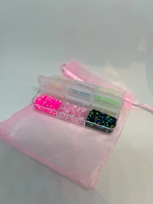 Bedezzel kit mini  - Säljer ett set med små, mixade plastpärlor i olika färger som neonrosa, blå, grön, svart och gul. Pärlorna kommer i en genomskinlig plastask med flera fack och en söt rosa organzapåse. Perfekt för att skapa egna smycken eller accessoarer.