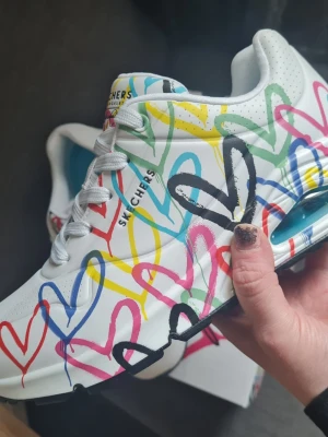 Helt nya vta Skechers sneakers med hjärtmönster - Färgglada Skechers sneakers i vitt med grafiska hjärtan i blått, rött, gult, grönt och svart över hela skon. Skorna har vit sula med svart detalj, luftbubbla i hälen och vita skosnören. Perfekt för dig som gillar statement sneakers och streetstyle. Helt nya och oanvända i kartong! Stl 37