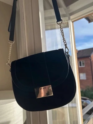 Svart axelväska från DKNY - Snygg svart axelväska från DKNY med rundad form och silverfärgad kedjedetalj på axelremmen. Väskan har ett lock med metallspänne där DKNY-loggan syns tydligt. Materialet är slätt och stilrent, perfekt för att lyfta vilken outfit som helst.