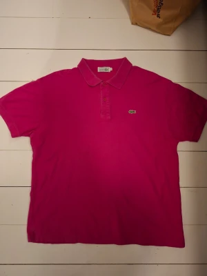 Rosa pikétröja från Lacoste - Säljer en klassisk rosa pikétröja från Lacoste med den ikoniska gröna krokodilloggan på bröstet. Tröjan har krage, knappar framtill och korta ärmar. Perfekt för dig som gillar stilrena och sportiga plagg. Finns viss slitage vid krage men annars bra.