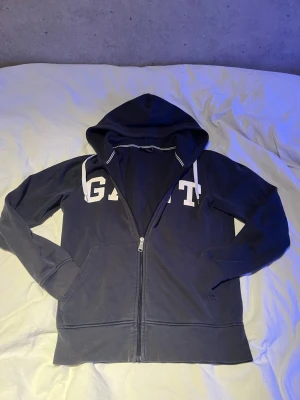 GANT zip tröja - Snygg mörkblå hoodie från GANT med vit logga över bröstet. Tröjan har dragkedja framtill, två fickor och huva med vita snören. Perfekt för en avslappnad och sportig stil.