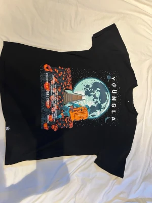 Svart YOUNGLA - Cool svart t-shirt från YOUNGLA med färgstarkt tryck på framsidan som visar en astronaut, måne och rymdtema. T-shirten har korta ärmar och rund hals. Materialet känns mjukt och skönt, perfekt för en avslappnad stil.