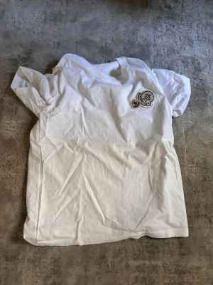  Vit Moncler t-shirt med logga - Säljer en klassisk vit t-shirt från Moncler med rund hals och korta ärmar. På bröstet sitter Monclers ikoniska broderade logga i vitt, rött och blått. T-shirten är gjord i mjuk bomull och har en clean, stilren look som passar till det mesta.