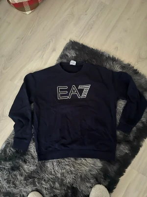 Mörkblå sweatshirt från EA7 Armani - Mörkblå sweatshirt från EA7 Emporio Armani med stor vit EA7-logga på bröstet. Klassisk rund halsringning och långa ärmar. Perfekt för en avslappnad och stilren look.