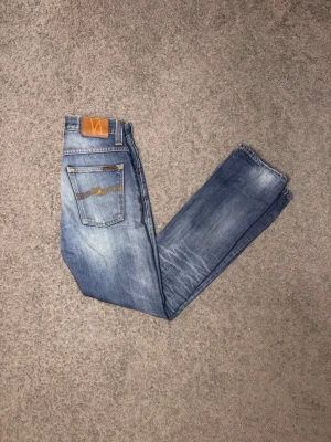Blå slitna jeans från Nudie Jeans - Säljer ett par blå jeans från Nudie Jeans med snyggt slitna detaljer och klassisk femficksmodell. Jeansen har raka ben och kontrastsömmar samt en brun patch bak i midjan. Perfekta för en avslappnad och trendig look.