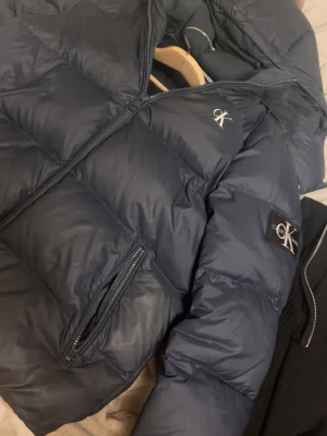 Mörkblå pufferjacka från Calvin Klein - Snygg mörkblå pufferjacka från Calvin Klein med dragkedja framtill, två sidofickor med dragkedja och CK-logga på bröstet och ärmen. Jackan har hög krage och quiltad design som ger en riktigt modern look. Perfekt för kalla vinterdagar.